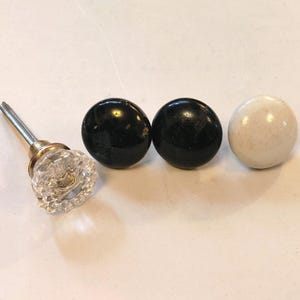 May include: Three cabinet knobs, two black and one white. The black knobs are round and smooth. The white knob is round and has a slightly textured surface. The knobs are all approximately 2.5 cm in diameter.