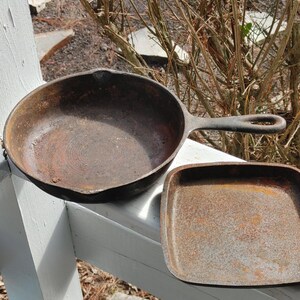 May include: Two cast iron skillets, one round and one square, both with a rusty brown patina.