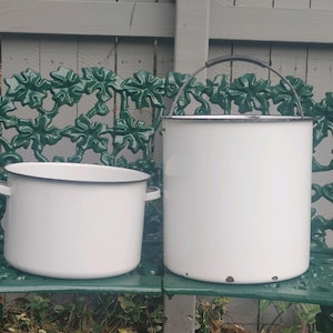 May include: Two white enamelware containers with black rims. The larger container has a black handle and is taller than the smaller container. Both containers are sitting on a green metal surface.