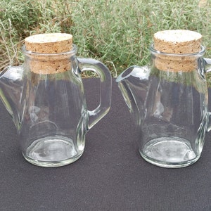 Könnte beinhalten: Zwei klare Glaskrüge mit Korkdeckeln. Die Krüge haben eine einzigartige Form mit einem Ausguss und einem Griff.