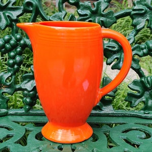 May include: A bright orange ceramic pitcher with a rounded body and a flared spout. The pitcher has a handle on the side and a wide base.