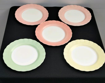 May include: Five pastel-colored ceramic dinner plates with scalloped edges. The plates are arranged in a circle on a black surface. The plates are white in the center and have a pastel pink, yellow, green, and light pink rim.