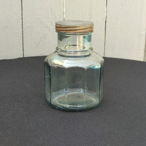 Peut inclure: Un pot en verre transparent avec un couvercle en métal. Le pot a une forme hexagonale et est posé sur une surface noire.