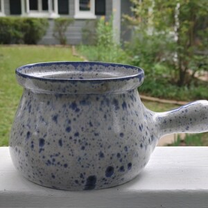 May include: A white ceramic soup bowl with a blue speckled glaze and a handle. The bowl is round and has a slight lip around the rim.