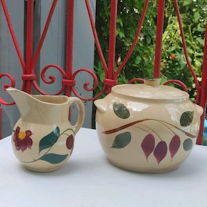 May include: A set of two vintage hand-painted pottery pieces. A cream-coloured pitcher with a green and burgundy floral design and a cream-coloured covered casserole dish with a green and burgundy floral design.
