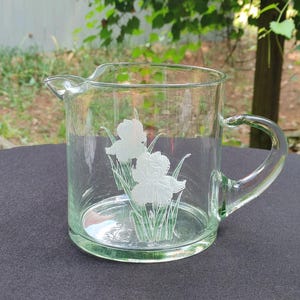 Könnte beinhalten: Eine klare Glaskanne mit einem weißen, geätzten Design von Irisblüten. Die Kanne hat einen Griff und einen Ausguss.