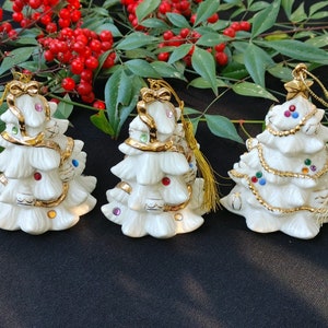May include: Three white porcelain Christmas tree ornaments with gold trim and colorful jewels. Each ornament has a gold tassel.