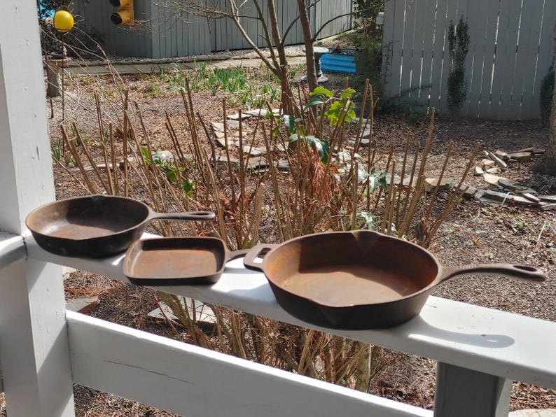 May include: Three cast iron skillets of different sizes, two round and one square, are lined up on a white wooden railing. The skillets are all rusty and have a worn look.