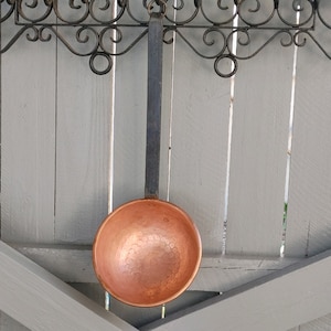 May include: A copper ladle with a hammered finish hangs from a wrought iron rack against a light gray wood background.