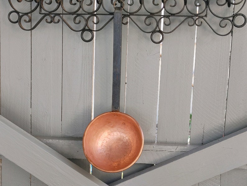 May include: A copper ladle with a hammered finish hangs from a wrought iron rack against a light gray wood background.