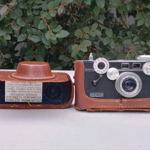 May include: A vintage silver and black 35mm film camera with a brown leather case. The camera has a silver lens with a black ring around it. The case has a label with the text "COLOR-MATIC SETTINGS RECOMMENDED FOR AVERAGE OUTDOOR CONDITIONS COLOR FILM-YELLOW SETTINGS BLACK & WHITE FILM-RED SETTINGS FLASH SYNCHRONIZES AT GREEN".