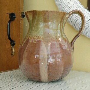 May include: A handcrafted ceramic pitcher with a bulbous body and a curved handle. The pitcher features a gradient of colors, transitioning from a light green at the top to a warm brown at the base. The pitcher is approximately 10 inches tall.