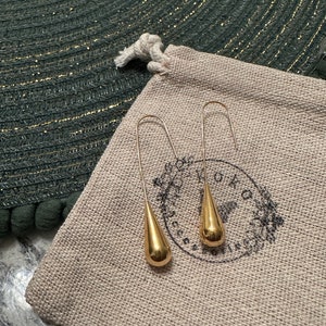 May include: A pair of gold teardrop earrings on a beige fabric pouch with a logo that says "OMO Jewellery".