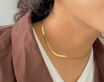 Op de afbeelding: Een gouden ketting met een delicaat, slangachtig ontwerp. De ketting wordt gedragen door een persoon met bruin haar en een bruin jack.