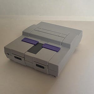 Op de afbeelding: Een grijze Super Nintendo Entertainment System (SNES) console. De console heeft een rechthoekige vorm met paarse knoppen en een zwart paneel. Het ontwerp is een klassieke retro gaming console.