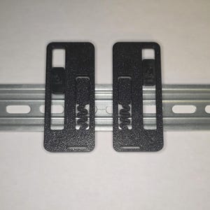 May include: Two black rectangular DIN rail clips with a textured surface are shown, mounted on a silver metal DIN rail. Each clip has a spring and a small black component. The background is plain white.