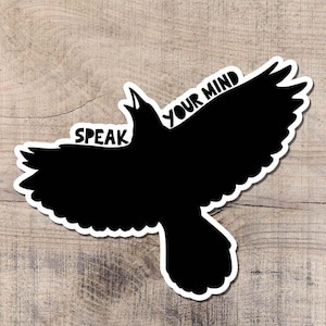 Puede incluir: Adhesivo de cuervo negro con las palabras "SPEAK YOUR MIND" en blanco. El cuervo es una silueta con las alas extendidas y un borde blanco, sobre un fondo de madera marrón claro.
