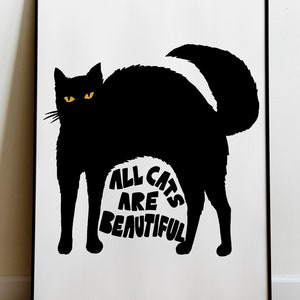 May include: Black cat silhouette art print with yellow eyes and the text "ALL CATS ARE BEAUTIFUL" on a white background. The artwork is framed in a black frame and displayed on a wooden floor.