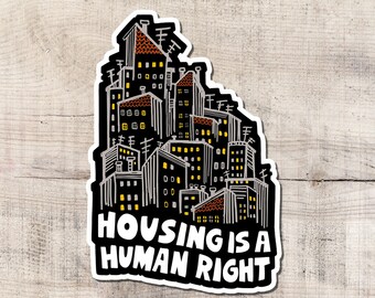 Puede incluir: Una pegatina con una ilustración estilizada de un paisaje urbano y el texto "HOUSING IS A HUMAN RIGHT". Los edificios están delineados en blanco sobre un fondo negro, con ventanas amarillas y tejados rojos.