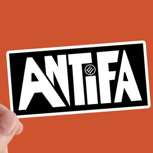 Peut inclure: Un autocollant rectangulaire avec le mot "ANTIFA" en lettres blanches grasses sur fond noir. Les lettres sont stylisées, avec un symbole au centre du "I". L'autocollant est tenu sur un fond orange.
