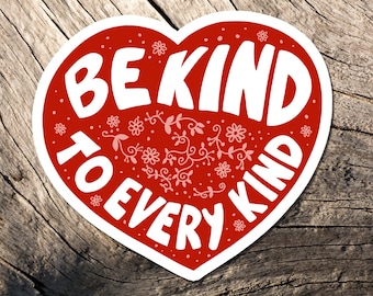 May include: A red heart-shaped sticker with the words "BE KIND TO EVERY KIND" in white lettering. The sticker has a white border and is decorated with floral and leaf designs. The sticker is placed on a weathered wooden surface.