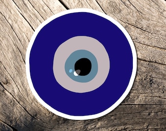 May include: A round sticker featuring a stylized blue eye design. The eye has a dark blue outer ring, a light blue iris, a gray ring, and a black pupil. The sticker is set against a weathered wood background.