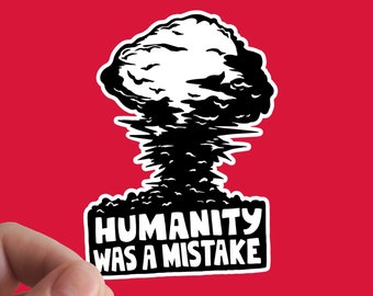 May include: A black and white sticker depicting a mushroom cloud explosion. The text "HUMANITY WAS A MISTAKE" is printed in white, bold font on a black banner. The sticker is held against a red background.