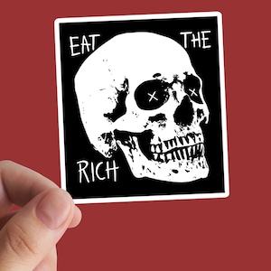 Può includere: Un adesivo in bianco e nero con un teschio e la scritta "EAT THE RICH". Il teschio ha gli occhi sbarrati. L'adesivo è tenuto in mano su sfondo rosso.