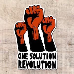 May include: A sticker featuring three raised fists in orange and black, with the text "ONE SOLUTION REVOLUTION" in bold, white letters. The sticker is set against a light wood grain background.