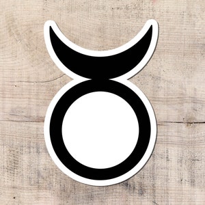 Puede incluir: Una pegatina en blanco y negro con el símbolo del zodiaco Tauro. El diseño incluye una forma de media luna sobre un círculo, todo delineado en negro con un interior blanco. La pegatina está colocada sobre una superficie de madera clara.