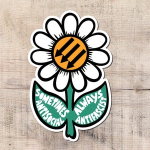 Puede incluir: Una pegatina de margarita blanca con un contorno negro, un centro naranja con tres flechas negras y hojas verdes. Las hojas tienen el texto "SOMETIMES ANTISOCIAL" y "ALWAYS ANTIFASCIST".