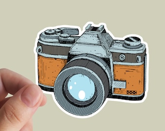 May include: A sticker featuring a vintage camera illustration. The camera has a large lens, a silver body, and orange accents. The design is a detailed line drawing with a retro aesthetic.