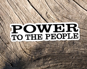 May include: A sticker with the words "POWER TO THE PEOPLE" in bold black letters on a white background. The sticker is placed on a weathered, textured wooden surface, creating a rustic contrast.