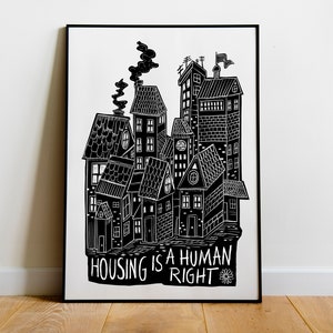 May include: Black and white art print depicting a collection of stylised buildings with chimneys and a flag. The text "HOUSING IS A HUMAN RIGHT" is at the bottom. The artwork is framed in a black frame.
