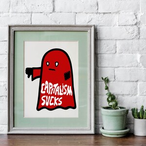 May include: Framed art print featuring a red ghost-like figure with a thumbs-down gesture. The text "CAPITALISM SUCKS" is written in white on the figure. The artwork is framed in a light green and gray frame.