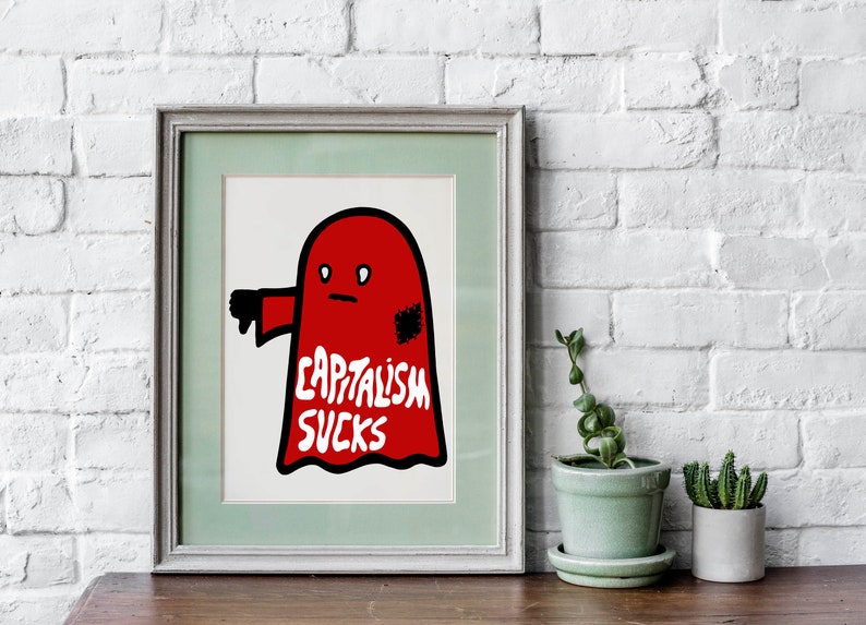 May include: Framed art print featuring a red ghost-like figure with a thumbs-down gesture. The text "CAPITALISM SUCKS" is written in white on the figure. The artwork is framed in a light green and gray frame.