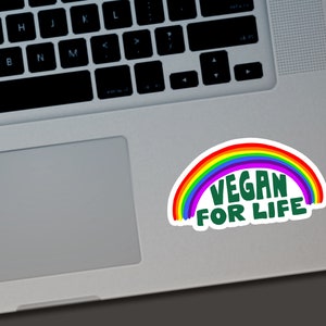 May include: A white sticker with a rainbow arc and the words "VEGAN FOR LIFE" in green. The sticker is on a laptop. The rainbow has red, orange, yellow, green, blue, and purple colors.