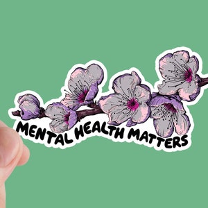 May include: A sticker featuring a branch of purple and white flowers with the text "MENTAL HEALTH MATTERS" in black. The sticker is held between two fingers against a light green background.