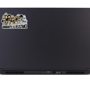 May include: A black laptop with a sticker featuring a cluster of brown and tan mushrooms. The sticker has the text "COMMUNITY IS RESISTANCE" in a white font with a black outline.