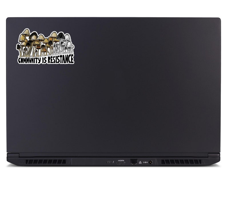 May include: A black laptop with a sticker featuring a cluster of brown and tan mushrooms. The sticker has the text "COMMUNITY IS RESISTANCE" in a white font with a black outline.