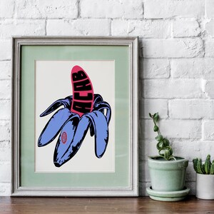 May include: Framed pop art print featuring a stylized banana with blue peels and a pink center. The word "ACAB" is printed in black on the banana. The artwork is displayed on a wooden surface with a white brick wall background.