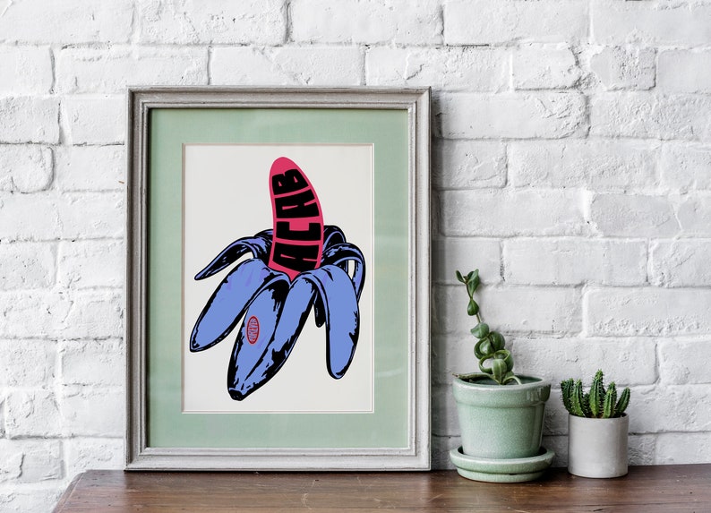 May include: Framed pop art print featuring a stylized banana with blue peels and a pink center. The word "ACAB" is printed in black on the banana. The artwork is displayed on a wooden surface with a white brick wall background.