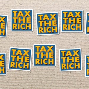 Op de afbeelding: Een verzameling stickers met de tekst "TAX THE RICH" in het geel, omlijnd met wit, op een blauwe achtergrond. De stickers liggen verspreid op een lichtbruin, getextureerd oppervlak.