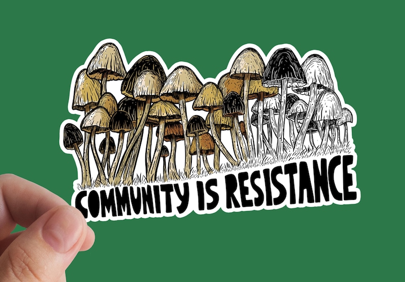 May include: A sticker featuring a cluster of detailed mushroom illustrations in shades of brown and black, with the text "COMMUNITY IS RESISTANCE" in bold, white letters. The sticker has a white border and is set against a green background.