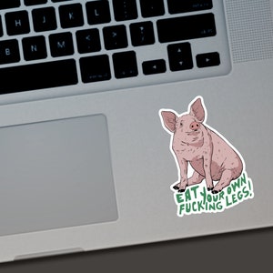 May include: A sticker featuring a cartoon pig in shades of pink and brown, sitting upright. The sticker has the text "EAT YOUR FUCKING LEGS!" in green. The sticker is placed on a laptop.