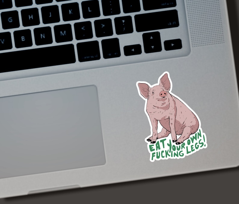 May include: A sticker featuring a cartoon pig in shades of pink and brown, sitting upright. The sticker has the text "EAT YOUR FUCKING LEGS!" in green. The sticker is placed on a laptop.