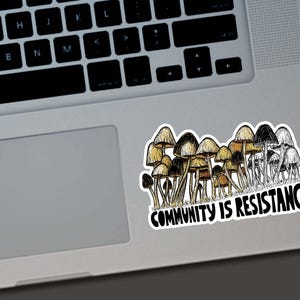May include: A sticker featuring a cluster of mushrooms in shades of brown, tan, and black. The sticker has the text "COMMUNITY IS RESISTANCE" in a bold, black font. The sticker is placed on a laptop.