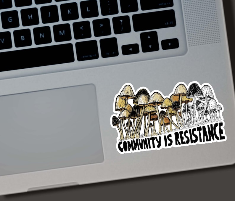 May include: A sticker featuring a cluster of mushrooms in shades of brown, tan, and black. The sticker has the text "COMMUNITY IS RESISTANCE" in a bold, black font. The sticker is placed on a laptop.