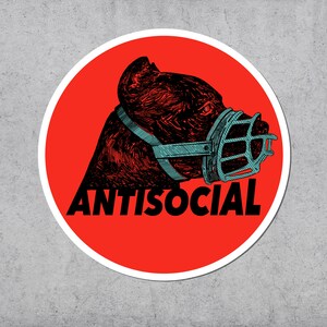 May include: A round sticker with a red background and a white border. The sticker features a detailed illustration of a dog's head wearing a blue muzzle. The word "ANTISOCIAL" is printed in black below the dog.