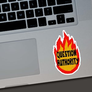 May include: A sticker with a flame design in red and yellow, with the words "QUESTION AUTHORITY" in black. The sticker is placed on a silver laptop.